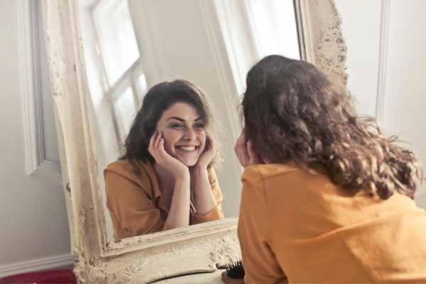 Mulher sorridente, vestindo uma blusa bege, apreciando seu reflexo frente ao espelho. Ela exibe uma expressão de felicidade, refletindo o autocuidado e a recuperação positiva após o término de um relacionamento.