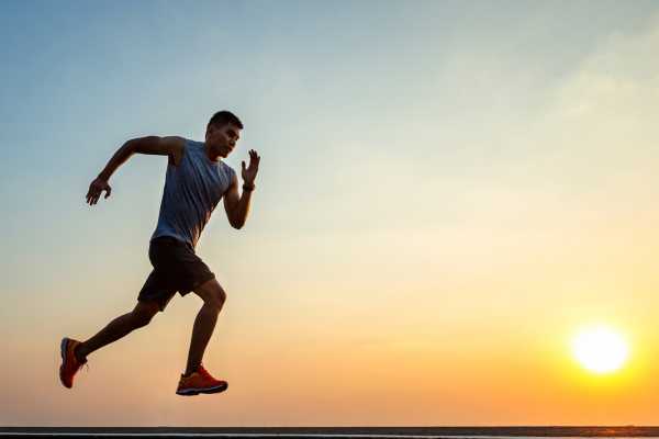 Na imagem, um homem está correndo em uma rua. Ele está vestindo Shorts escuros e um par de tênis de corrida. O homem está correndo pela estrada com o sol se pondo ao fundo. A cena captura a resiliência e o foco do homem enquanto ele se esforça para alcançar seus objetivos de condicionamento físico. A imagem transmite a mensagem da importância de manter um estilo de vida saudável e buscar metas pessoais.