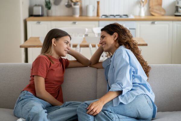 É a foto de uma mãe sentada em um sofá, apoiada com o braço direito no encosto, virada para uma menina sentada à sua direita, que é a sua filha. A mãe dirige um olhar de compreensão para a ela. E a filha está sorrindo, sentada virada para a mãe, apoiada com seu braço esquerdo sobre o encosto do sofá. Transmite algo muito bonito entre a mãe e sua filha, que deve ser uma pré-adolescente. A mãe está envolvendo a filha e há uma atmosfera de intimidade e compreensão, entre elas.