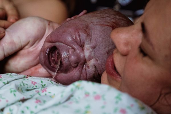 É a foto de uma mãe que acabou de receber o seu bebê, logo após o parto. Ela está deitada e o bebê abraçado por ela. É possível ver o rosto dele totalmente roxo e parte de seu tórax. A sua expressão é de muita dor e sofrimento, pois como é feito o parto normal, o cordão umbilical é cortado quando ainda está pulsando e o bebê fica privado de oxigênio por um tempo.