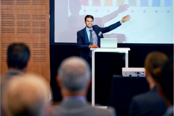 É a foto de um executivo fazendo uma apresentação em uma empresa, com a expressão sorridente e um gestual amplo, mostrando espontaneidade em vez de medo de falar em público. E essa é uma das 4 condições necessárias para se exercer influência quando se fala em público.