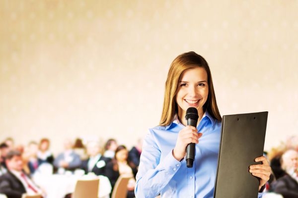 É a foto de uma moça com um microfone em uma mão e com a outra segurando um notebook. Está sorridente e com uma expressão de simpatia, e atrás aparece um público, provavelmente, formado por executivos de alguma empresa. Para quem está falando em público, ela parece tranquila, de bom humor e capaz de expressar emoções relacionadas com o que fala. E isso é a chave para se comunicar exercendo persuasão.